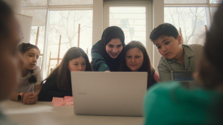 Rahaf cercada de crianças em uma mesa apontando para a tela de um notebook Samsung durante um de seus workshops