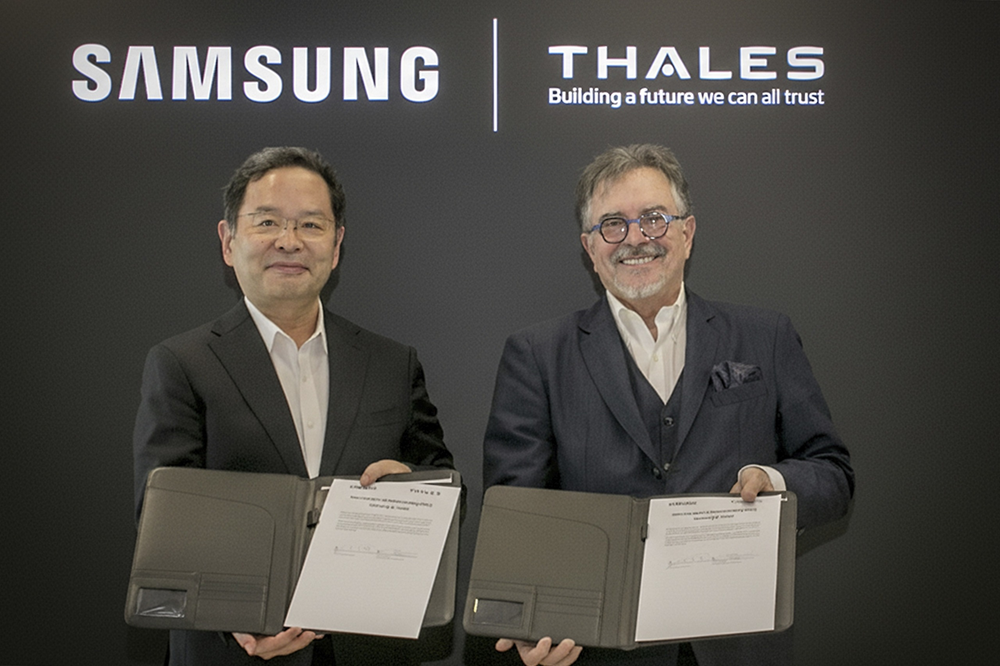 Representantes da Samsung e da Thales apertam as mãos para firmar acordo em frente à um painel escuro com os logotipos de ambas as empresas
