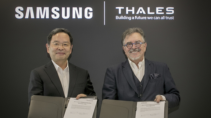 Representantes da Samsung e da Thales apertam as mãos para firmar acordo em frente à um painel escuro com os logotipos de ambas as empresas