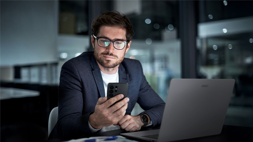 Homem usando smartphone, notebook e smartwatch Samsung em ambiente corporativo