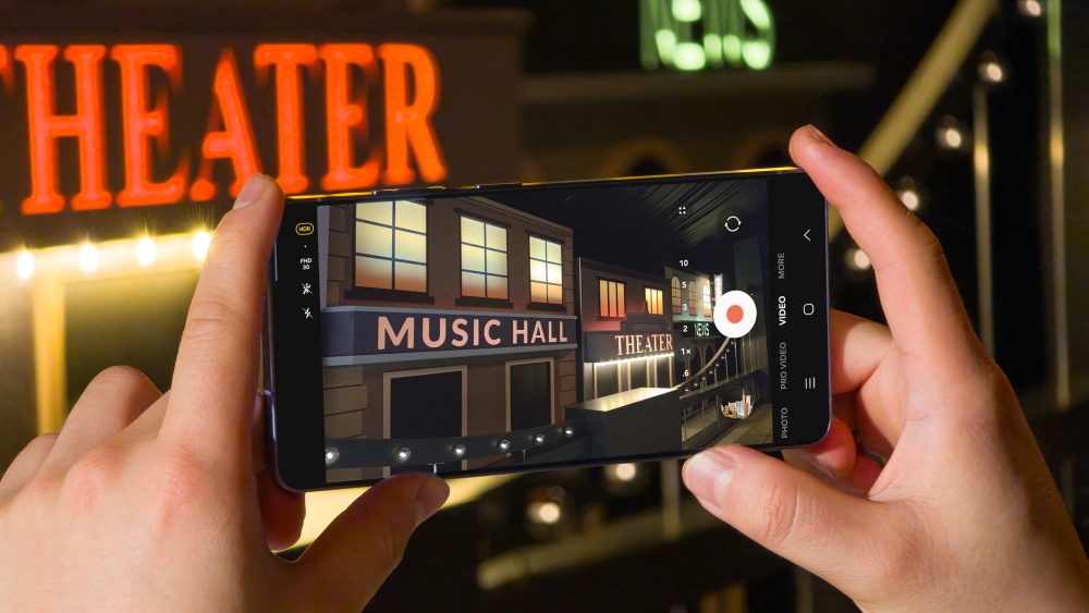 Um celular segurado pelas mãos apontando para a fachada de um teatro. O teatro possui três janelas iluminadas e a frase "Music Hall" abaixo, além da palavra "Theater" em vermelho.