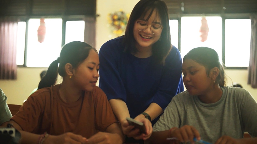 Brigitta Gunawan entre duas jovens mostrando a tela de um smartphone Galaxy