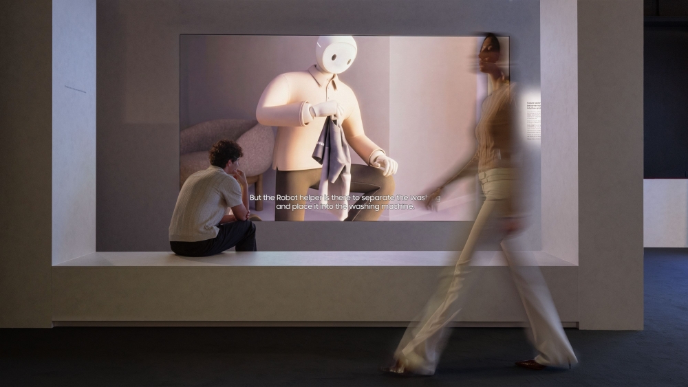 Foto da zona 'A Glimpse of Tomorrow', da exposição 'Design is an act os Love", mostra um homem sentado frente à uma tela com um robô enquanto uma mulher caminha pelo espaço.