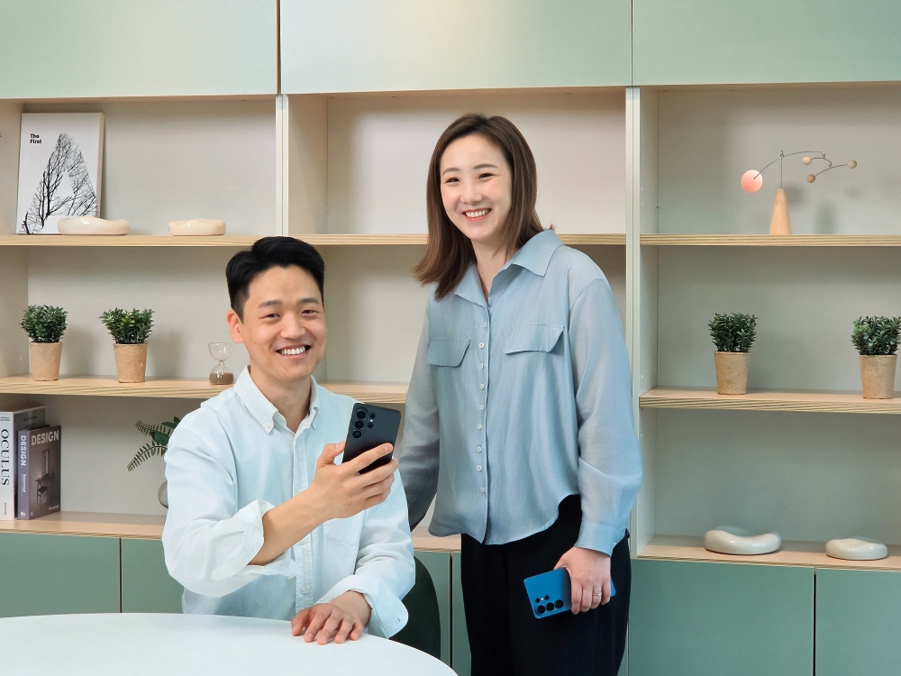 A imagem mostra os desenvolvedores Junseang Min e Sunmi Yoo, da divisão Mobile Experience da Samsung Electronics. Junseang está à esquerda da foto, sentado, usando uma camisa social azul clara e segurando um celular na mão direita. Ele também está sorrindo. Sunmi Yoo, à direita, está de pé, também sorrindo, segurando um celular na mão esquerda e usando uma camisa social azul um pouco mais escura e calça preta. Ao fundo, móveis com prateleiras de decoração, incluindo pequenos vasos com plantas e livros.