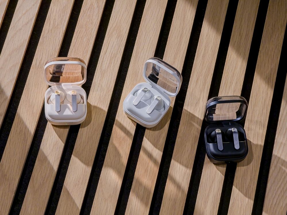 A imagem mostra três unidades do Samsung Galaxy Buds4 Pro, nas cores rosé, branca e preta. Os fones, dentro de suas cases, aparecem em cima de uma superfície de madeira, e estão iluminados pela luz solar.