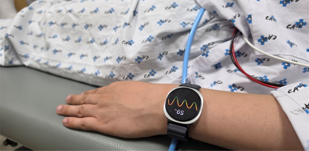 A imagem mostra uma pessoa deitada, com roupa hospitalar, mas o foco está no braço dela vestindo um relógio no pulso. O relógio mostra a porcentagem "59%", e o desenho do que parece ser um medidor de frequência cardíaca. Além disso, fios aparecem por cima da roupa da pessoa deitada sobre uma maca cinza.