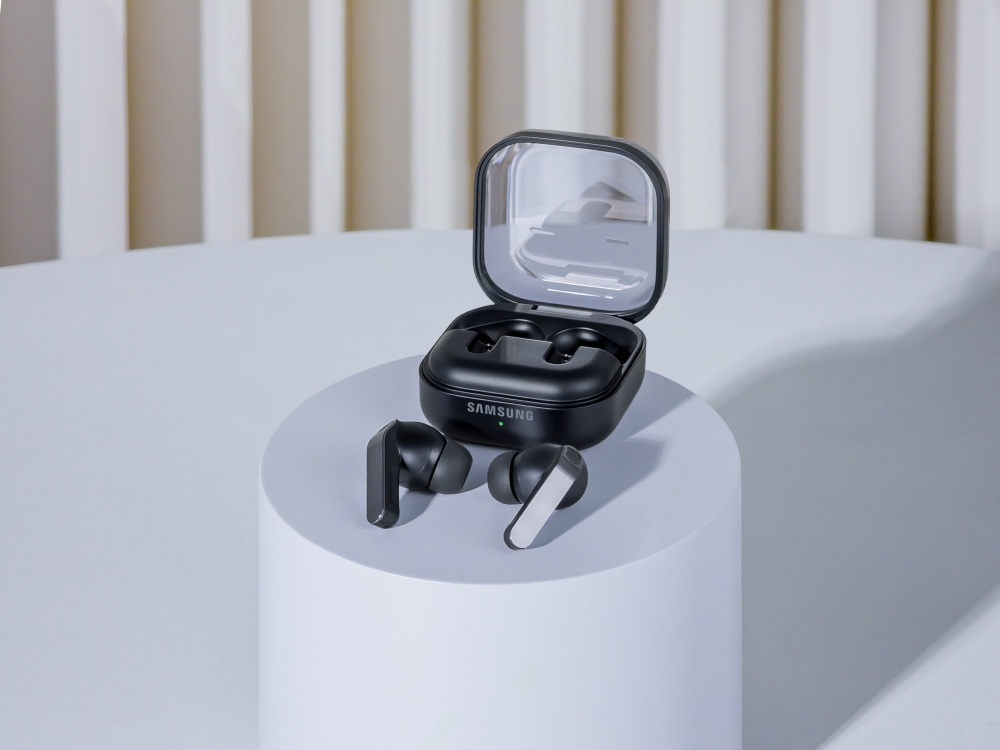 A imagem mostra uma unidade do fone de ouvido Samsung Galaxy Buds4 Pro, na cor preta. Os fones aparecem fora do estojo de carregamento, e estão em cima de uma pequena superfície circular, que por sua vez parece estar em cima de uma mesa - ambas as superfícies em um tom de cor acinzentado.