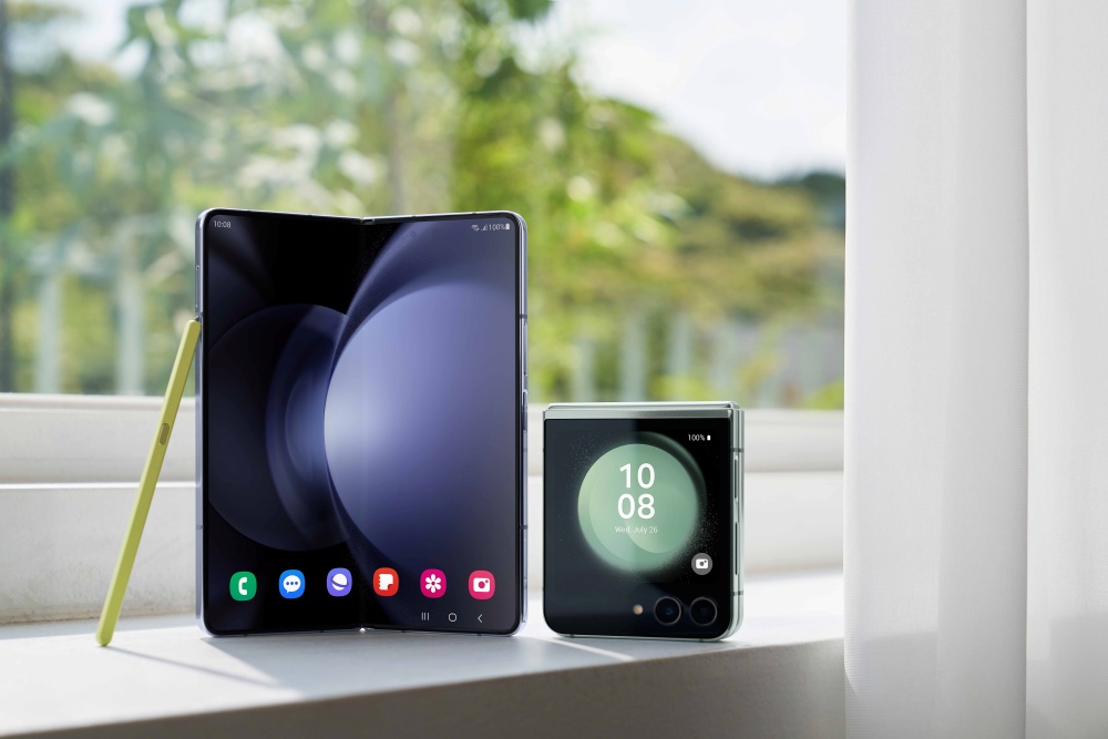 A imagem mostra dois celulares apoiados sobre uma superfície branca em frente a uma janela. Os celulares são Galaxy Z Fold5, que aparece semi-aberto em pé e, ao lado esquerdo, uma caneta S Pen amarela apoiada no aparelho. Ao lado dele, um Galaxy Z Flip5 fechado com a tela externa ligada. Uma cortina branca está mais para a direita da imagem, e o fundo da janela mostra um fundo verde com árvores.