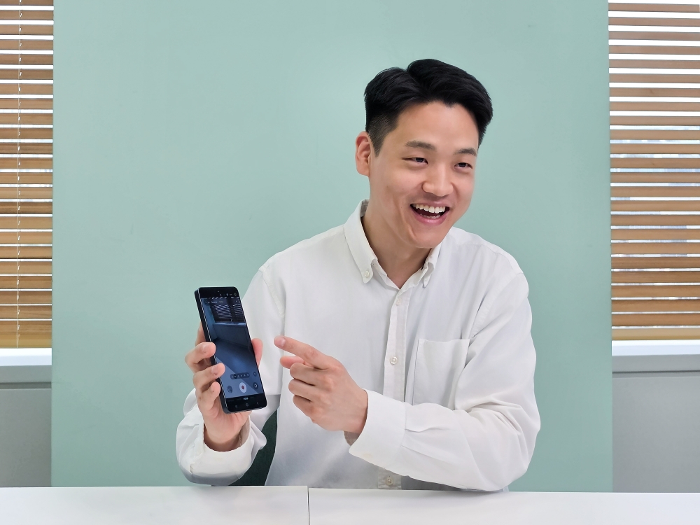 O desenvolvedor Junseang Min, da divisão Mobile Experience da Samsung Electronics. Junseang aparece usando uma camisa social branca, sentado, segurando um celular com a mão direita e apontando para ele com a mão esquerda. A mesa à frente dele é branca, e a parede ao fundo é em um tom de verde oliva.