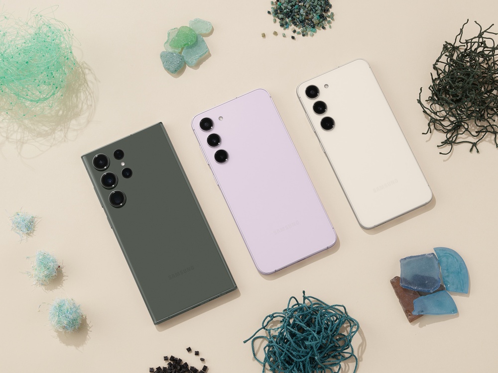 A imagem mostra três celulares, um ao lado do outro, em cima de uma bancada com vários outros objetos (pedras, fiapos coloridos). Os smartphones da linha Galaxy S23 aparecem em três modelos e cores diferentes: da esquerda para a direita, são eles o Galaxy S23 Ultra na cor grafite; o Galaxy S23+ na cor violeta; e o Galaxy S23 na cor creme.
