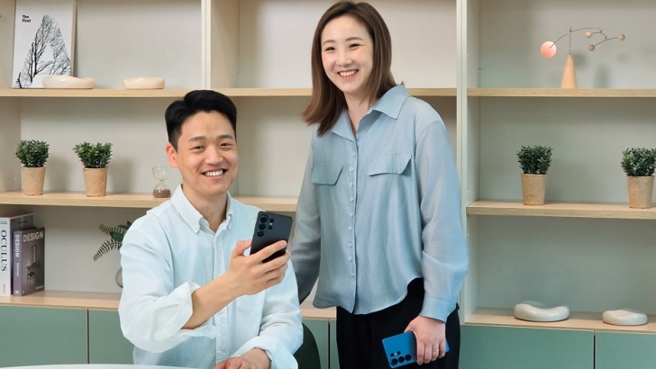 A imagem mostra os desenvolvedores Junseang Min e Sunmi Yoo, da divisão Mobile Experience da Samsung Electronics. Junseang está à esquerda da foto, sentado, usando uma camisa social azul clara e segurando um celular na mão direita. Ele também está sorrindo. Sunmi Yoo, à direita, está de pé, também sorrindo, segurando um celular na mão esquerda e usando uma camisa social azul um pouco mais escura e calça preta. Ao fundo, móveis com prateleiras de decoração, incluindo pequenos vasos com plantas e livros.