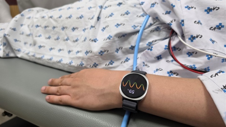 A imagem mostra uma pessoa deitada, com roupa hospitalar, mas o foco está no braço dela vestindo um relógio no pulso. O relógio mostra a porcentagem "59%", e o desenho do que parece ser um medidor de frequência cardíaca. Além disso, fios aparecem por cima da roupa da pessoa deitada sobre uma maca cinza.