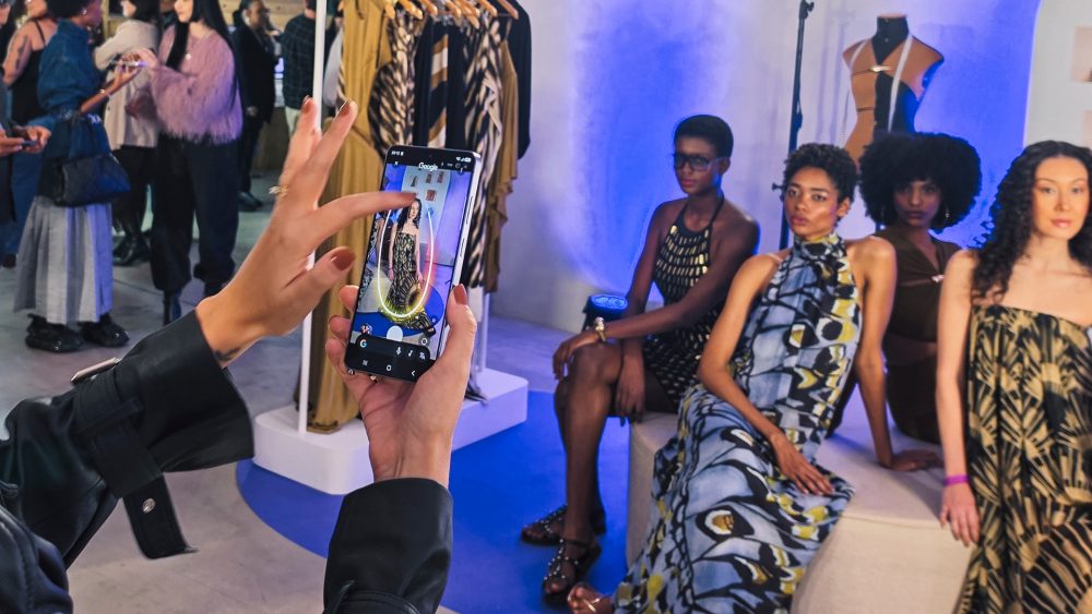 Pessoa segura um Galaxy S26 Ultra e utiliza o recurso Circule para Pesquisar no Google para destacar o vestido de uma modelo exibida na tela do smartphone. Ao fundo, modelos vestindo diferentes looks participam de uma apresentação de moda, enquanto araras de roupas e convidados compõem o ambiente do Galaxy S26 Ultra Fashion Show.