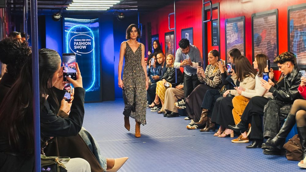 Modelo desfila por uma passarela cercada por convidados durante o Galaxy S26 Ultra Fashion Show. Pessoas sentadas dos dois lados registram o momento com seus smartphones, enquanto ao fundo uma estrutura iluminada exibe a identidade visual do evento.