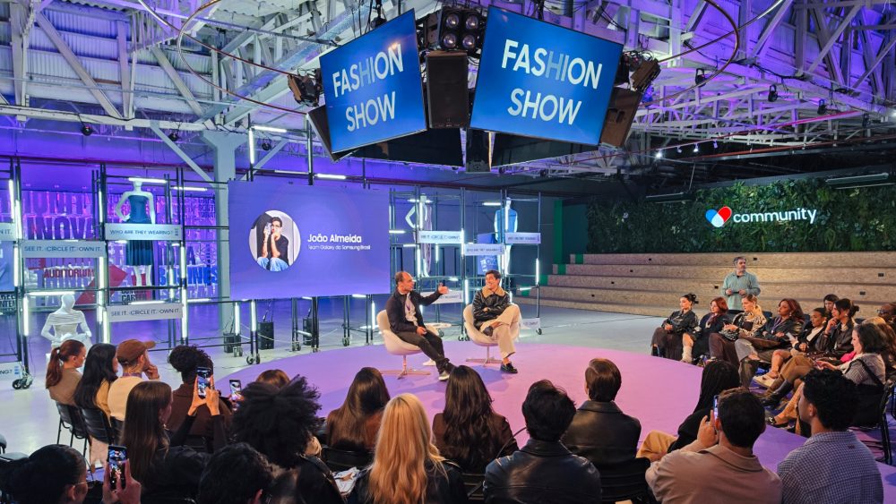 Renato Citrini, gerente sênior de produto de Mobile Experience da Samsung Brasil, e o Team Galaxy João Almeida participam de uma conversa no palco durante o Galaxy S26 Ultra Fashion Show. Os dois estão sentados em poltronas no centro do espaço, cercados por convidados. Ao fundo, um telão exibe a foto e o nome de João Almeida, enquanto estruturas iluminadas e elementos cenográficos do evento compõem o ambiente.