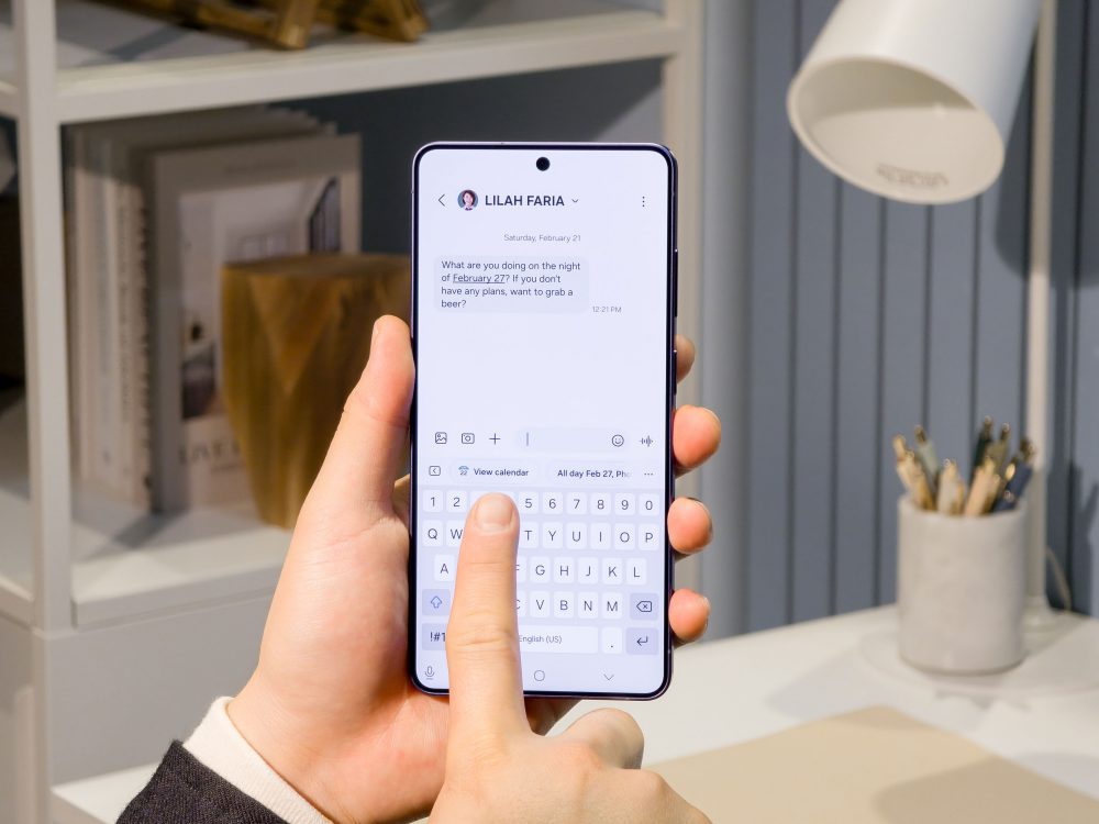 Pessoa segurando smartphone Galaxy S26 Ultra com conversa de mensagem aberta, mostrando texto e teclado virtual. Ambiente interno com estante, luminária e porta-lápis ao fundo, sugerindo contexto de trabalho ou estudo.
