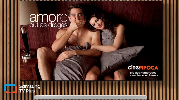 Imagem promocional do serviço Samsung TV Plus mostra uma TV Samsung, posicionada em um acabamento ripado de madeira, com cena do filme "Amor e outras drogas", disponível no canal Cine Pipoca, um dos canais do serviço gratuito, como parte da programação especial do serviço gratuito para o Dia dos Namorados.
