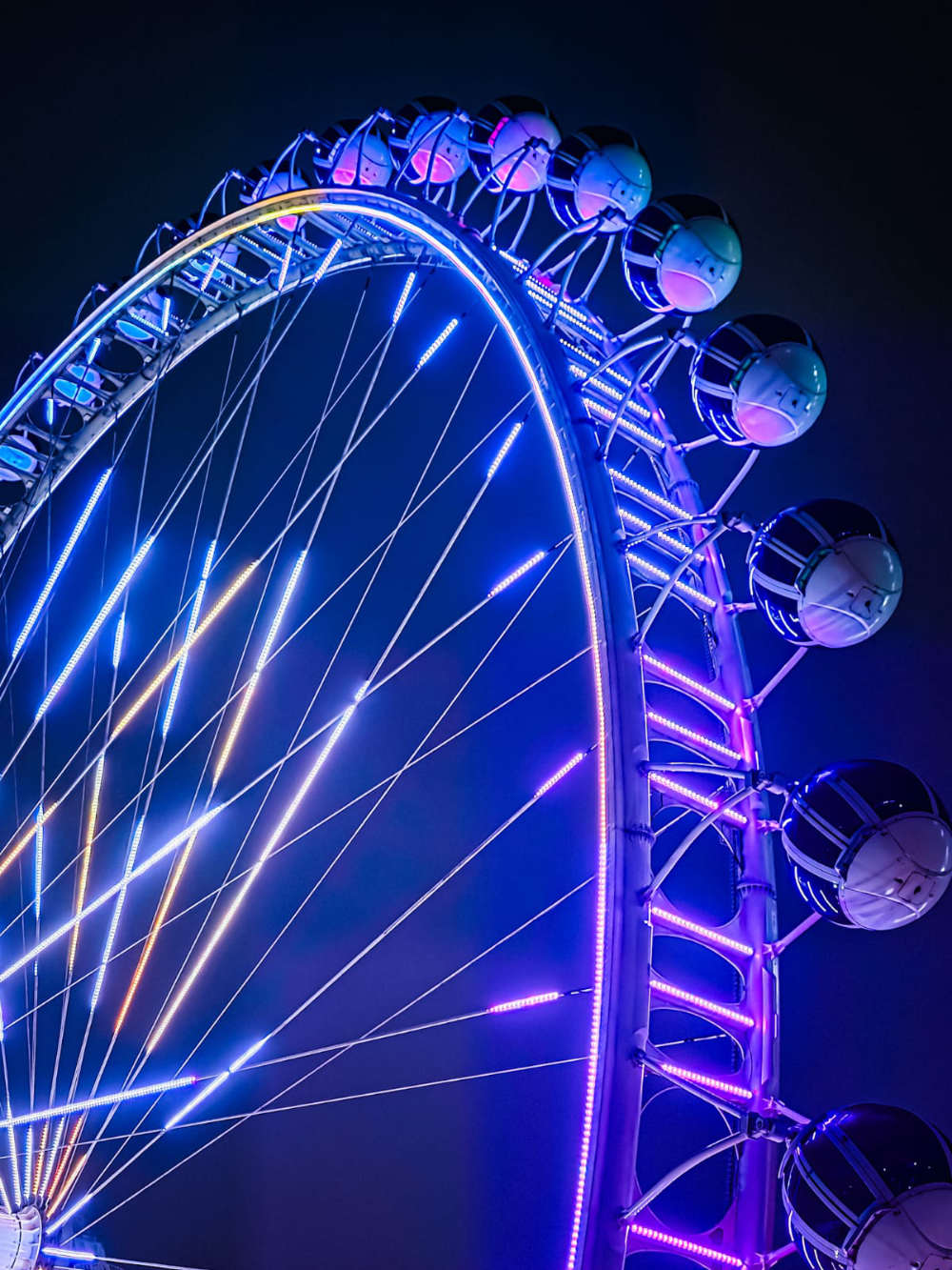 Close da Roda-Gigante Galaxy iluminada por luzes LED azuis e roxas contra o céu noturno, destacando estrutura e cabines do brinquedo.