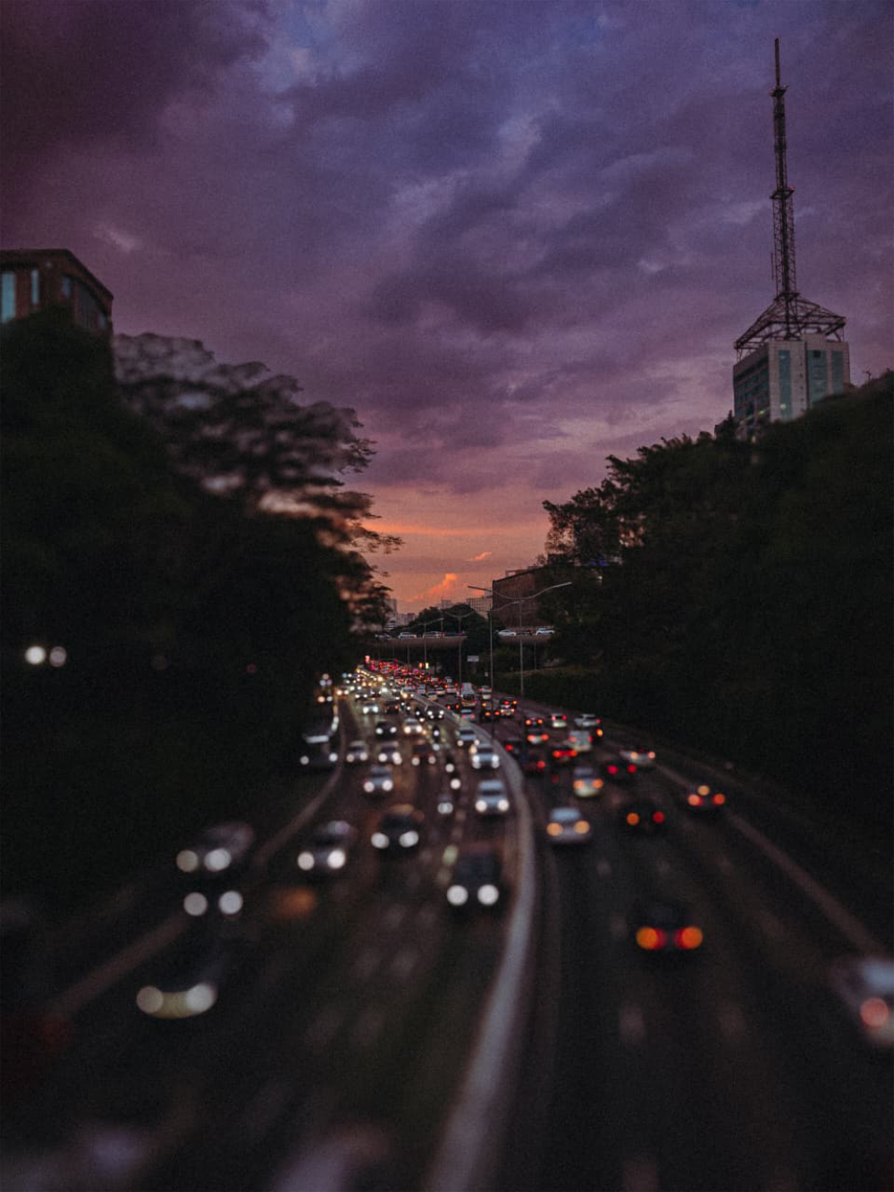 Vista elevada da Avenida 23 de Maio, em São Paulo, movimentada ao entardecer, com veículos em circulação e céu com tons arroxeados ao fundo.
