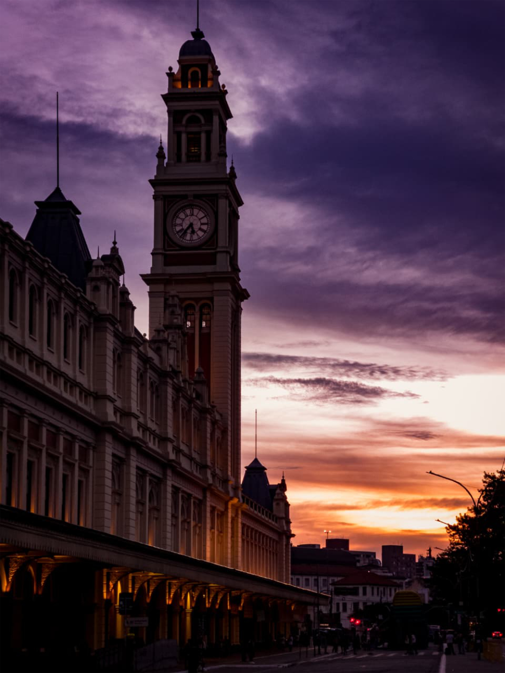 Fotografia da Estação da Luz, em São Paulo, ao pôr do sol, com a torre do relógio em destaque sob um céu em tons de roxo e laranja e parte da fachada histórica iluminada.