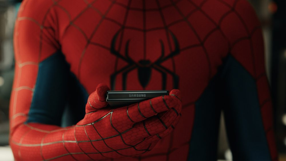 Close do torso do Homem-Aranha segurando um smartphone Samsung Galaxy Z Flip dobrado na altura do peito, com o logotipo Samsung visível na dobradiça. O personagem observa a tela externa do dispositivo.