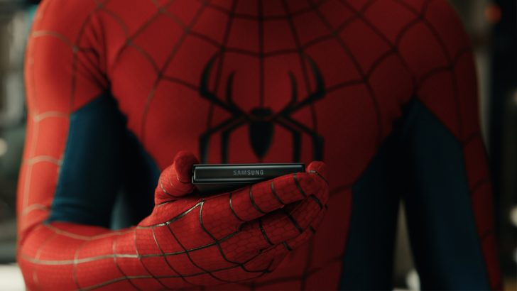 Close do torso do Homem-Aranha segurando um smartphone Samsung Galaxy Z Flip dobrado na altura do peito, com o logotipo Samsung visível na dobradiça. O personagem observa a tela externa do dispositivo.