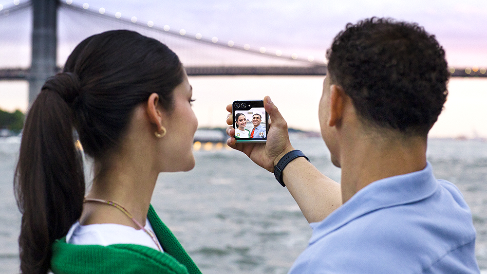 Uma imagem que mostra uma mulher e um homem, em frente a uma ponte e um lago. O homem segura um Galaxy Z Flip5 para tirar uma selfie dos dois.
