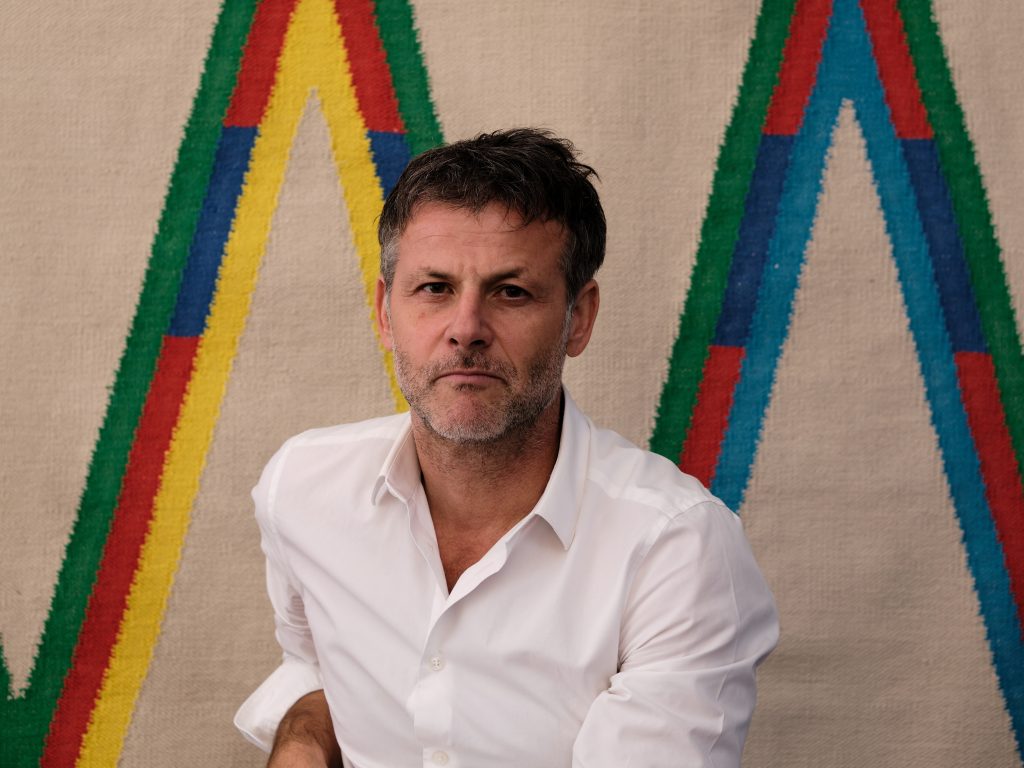 A imagem mostra o artista contemporâneo suíço Karim Noureldin em um retrato, vestindo uma camisa azul e posicionado em frente a um fundo claro.