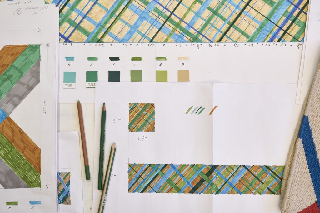 A imagem mostra estudos de composição desenvolvidos por Karim Noureldin sobre folhas de papel. Os desenhos apresentam padrões geométricos formados por linhas em tons de azul, verde e ocre, acompanhados de amostras de cores, anotações e lápis utilizados pelo artista durante o processo criativo.