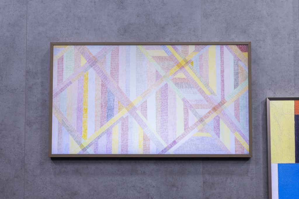 A imagem mostra uma TV Samsung The Frame exibindo uma obra abstrata de Karim Noureldin. A composição apresenta linhas diagonais e verticais em tons suaves de rosa, amarelo, azul e verde, instalada em uma parede com acabamento em concreto ao lado de outra obra de arte.