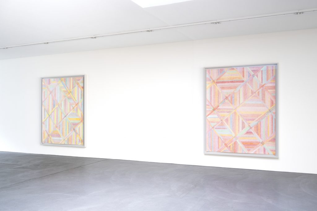 A imagem mostra duas obras abstratas de Karim Noureldin expostas em uma galeria de arte. As composições apresentam linhas geométricas e sobreposições de formas em tons suaves de rosa, amarelo, azul e verde, instaladas em uma parede branca.