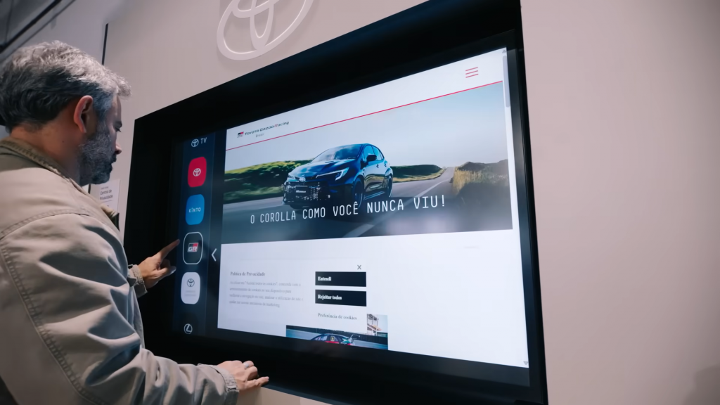 Pessoa utilizando uma tela profissional Samsung instalada em uma concessionária Toyota. O display exibe o portal da Toyota Gazoo Racing e um menu lateral com aplicativos da marca, demonstrando a solução de sinalização digital desenvolvida pela Samsung para modernizar a experiência dos clientes e otimizar a comunicação nas concessionárias.