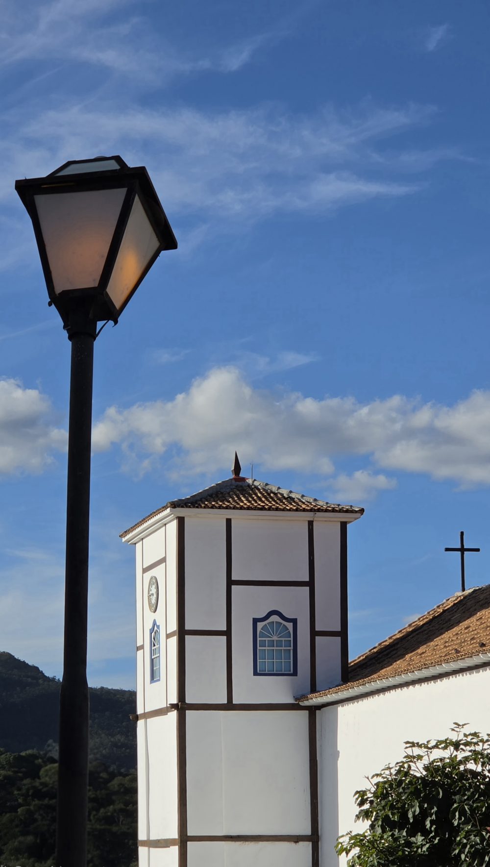 Vista da Igreja Matriz de Nossa Senhora do Rosário, em Pirenópolis (GO), com destaque para a torre branca de detalhes escuros e janelas azuis. Em primeiro plano, um poste de iluminação, enquanto ao fundo aparecem um céu azul com nuvens, morros e a cruz sobre o telhado da igreja.