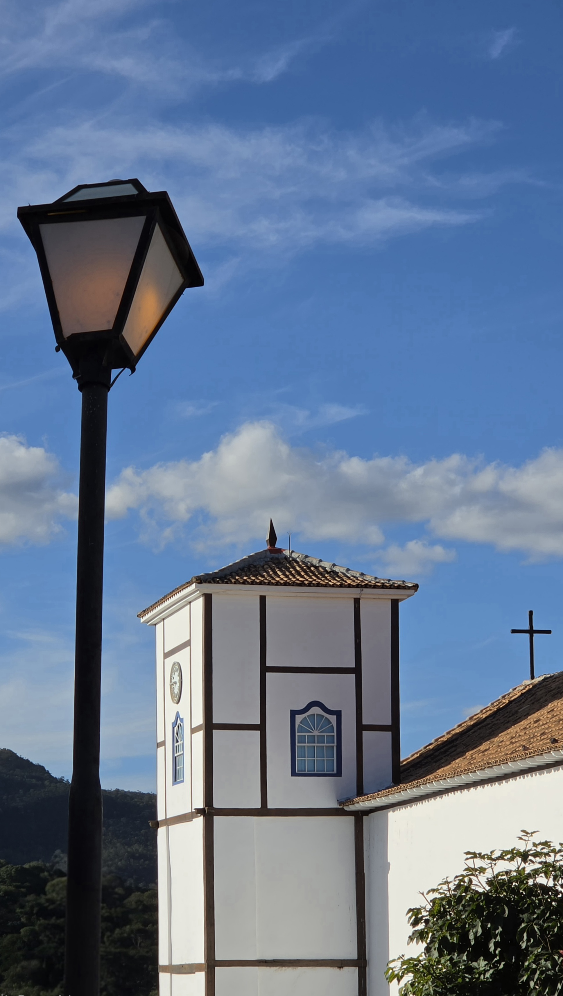 Vista da Igreja Matriz de Nossa Senhora do Rosário, em Pirenópolis (GO), com destaque para a torre branca de detalhes escuros e janelas azuis. Em primeiro plano, um poste de iluminação, enquanto ao fundo aparecem um céu azul com nuvens, morros e a cruz sobre o telhado da igreja.