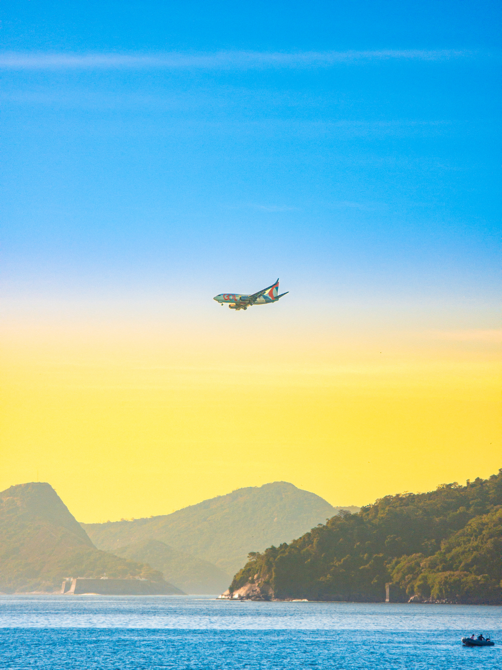 Avião comercial voa sobre a Baía de Guanabara durante o pôr do sol. Ao fundo, montanhas e ilhas compõem a paisagem, enquanto o céu apresenta um degradê entre tons de azul e dourado.
