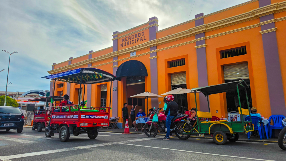 Fachada do Mercado Municipal de Parintins pintada em tons de laranja e roxo. Em frente ao prédio, motos, triciclos, carros e pessoas movimentam a rua.