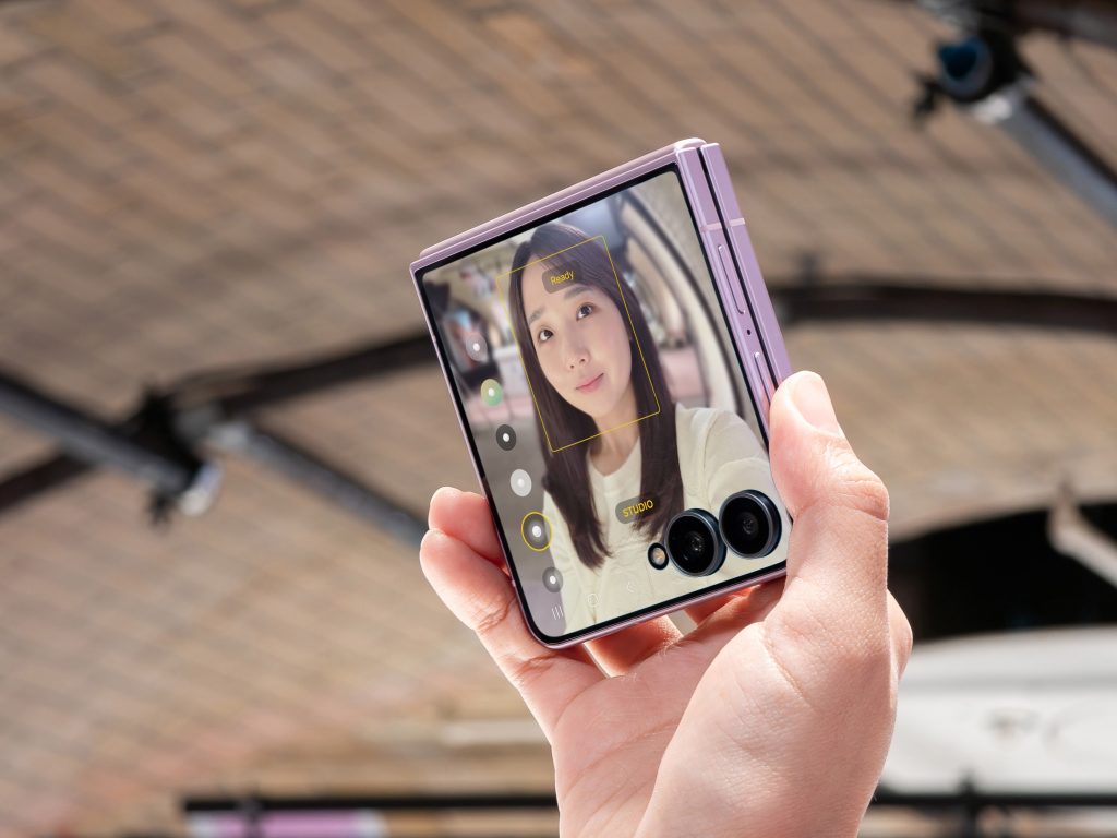 Fotografia mostra mão segurando o Galaxy Z Flip8 na cor Rosa dobrado. A tela externa, aberta com o aplicativo Câmera, mostra o rosto de mulher em foco. 
