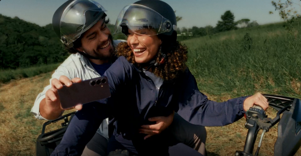 Um homem e uma mulher em cima de uma moto. Os dois estão sorrindo, usando capacete. O homem segura um Samsung Galaxy S26 com a mão direita, filmando os dois na moto.