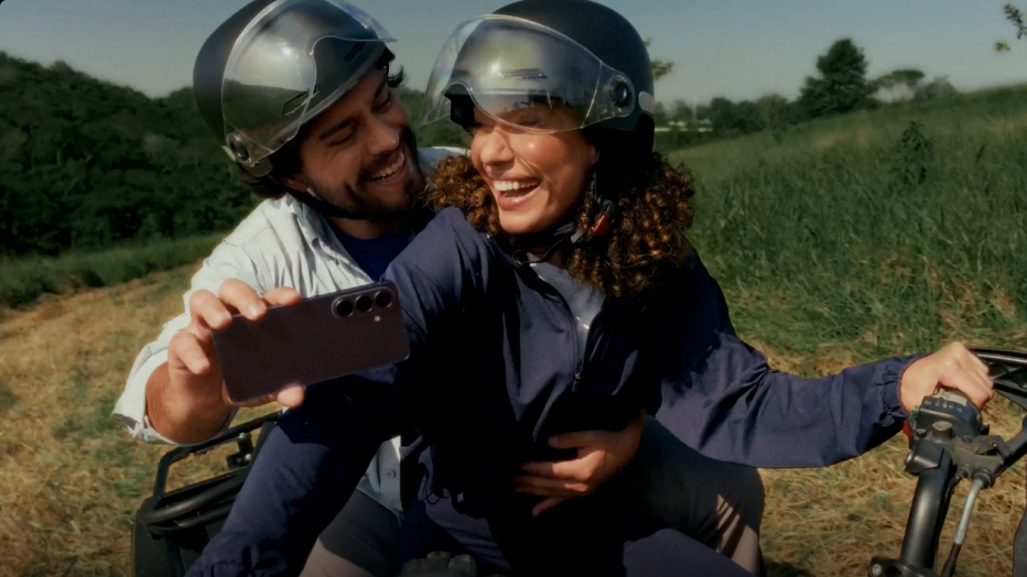 Um homem e uma mulher em cima de uma moto. Os dois estão sorrindo, usando capacete. O homem segura um Samsung Galaxy S26 com a mão direita, filmando os dois na moto.