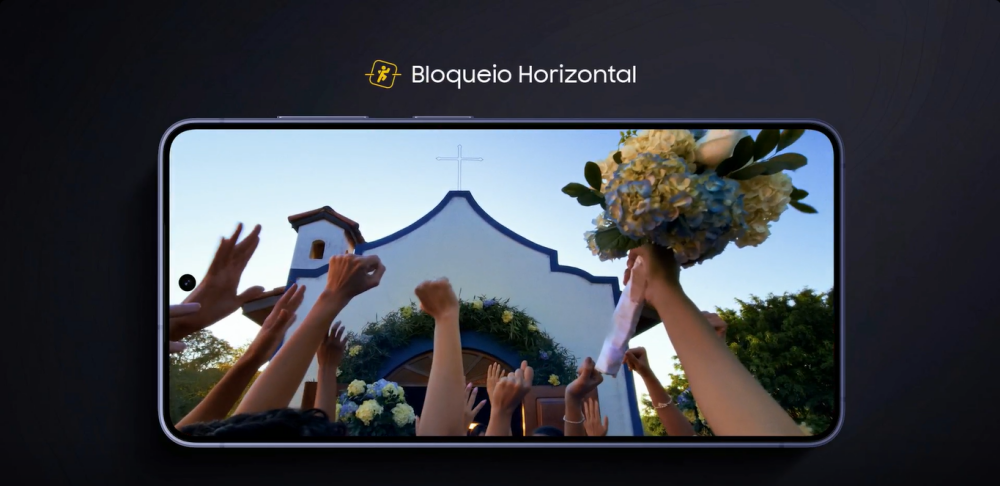 Um celular da linha Samsung Galaxy S26 deitado, com a tela ligada, usando a funçao de Bloqueio Horizontal durante a gravação de um vídeo. No vídeo da tela, várias mãos levantadas para cima tentando pegar um buque de flores.