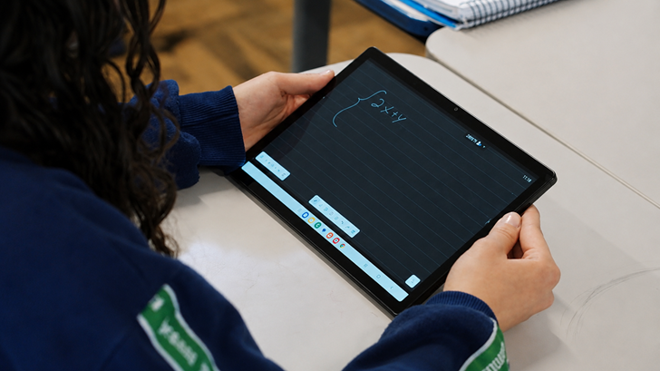 Vista aproximada de uma estudante sentada em uma sala de aula, segurando um tablet da Samsung na posição horizontal. Na tela do dispositivo, aparece uma atividade de matemática com uma equação escrita digitalmente. A cena destaca o uso de recursos tecnológicos no ambiente escolar, evidenciando como tablets e soluções digitais contribuem para tornar o aprendizado mais interativo, conectado e acessível.