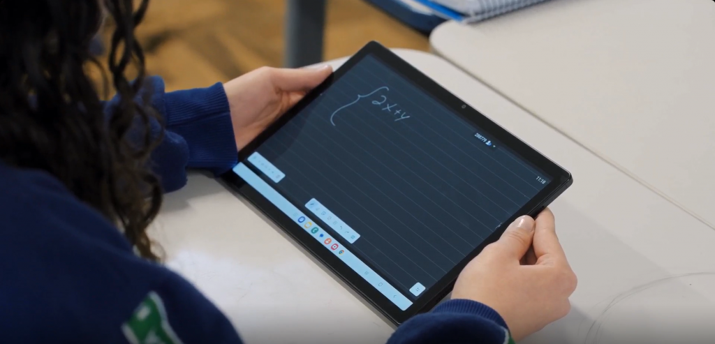 Vista aproximada de uma estudante sentada em uma sala de aula, segurando um tablet da Samsung na posição horizontal. Na tela do dispositivo, aparece uma atividade de matemática com uma equação escrita digitalmente. A cena destaca o uso de recursos tecnológicos no ambiente escolar, evidenciando como tablets e soluções digitais contribuem para tornar o aprendizado mais interativo, conectado e acessível.