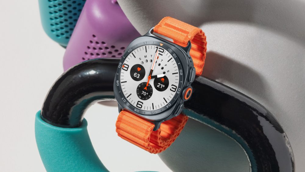 Galaxy Watch Ultra2 em Titânio Prata com mostrador analógico branco e Pulseira Trail Laranja apoiado sobre um kettlebell preto. Ao fundo, há kettlebells desfocados nas cores verde-água e roxo.