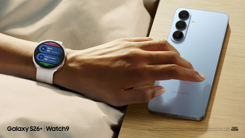 Pessoa usa um Galaxy Watch9 enquanto segura um Galaxy S26+ azul sobre uma mesa. Na tela do relógio, são exibidas as pontuações de Sono e Saúde do Coração integradas ao Samsung Health.
