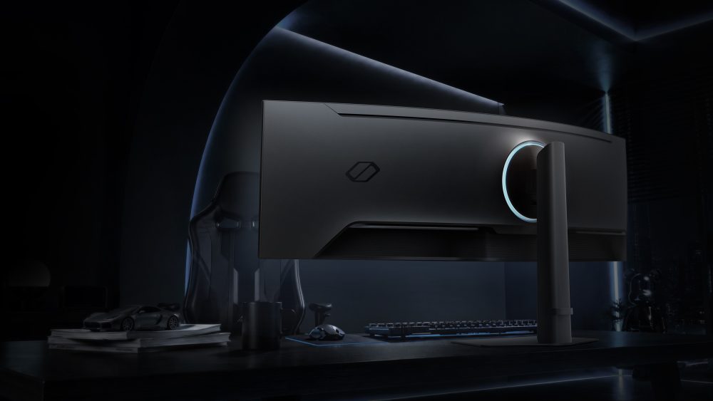 Vista traseira do monitor gamer Samsung Odyssey OLED G9 em um setup escuro, com iluminação ambiente, teclado, mouse e cadeira gamer sobre a mesa.
