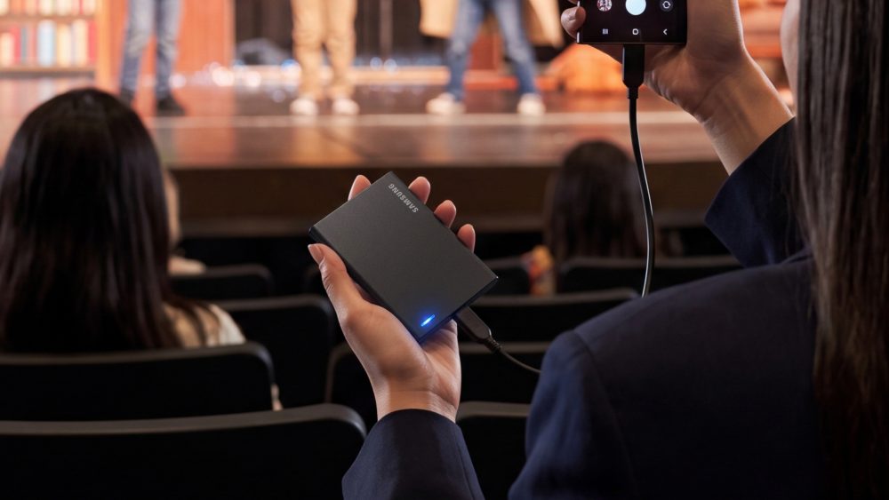 Pessoa utiliza um SSD portátil Samsung P7 conectado por cabo a um smartphone durante a gravação de uma apresentação, com o dispositivo apoiado em uma das mãos.