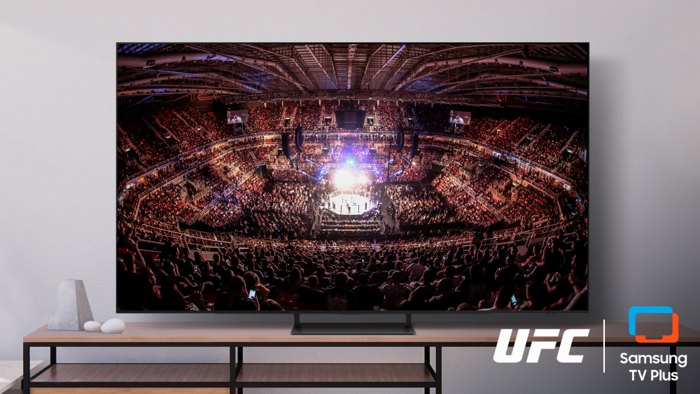 Televisão exibindo uma transmissão de um evento do UFC em uma arena lotada, com o octógono iluminado no centro e o público ocupando as arquibancadas. O logotipo do UFC aparece no canto inferior direito da imagem, ao lado do logotipo do Samsung TV Plus.
