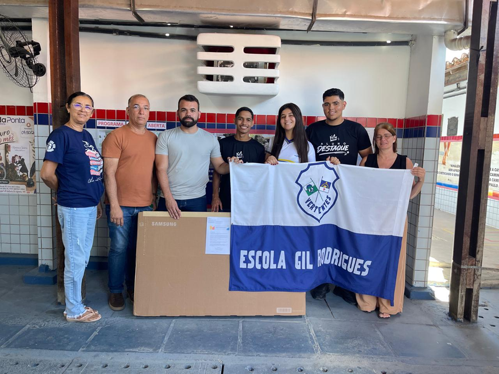 Grupo de seis pessoas posa para foto segurando uma bandeira azul e branca com o nome da Escola Gil Rodrigues