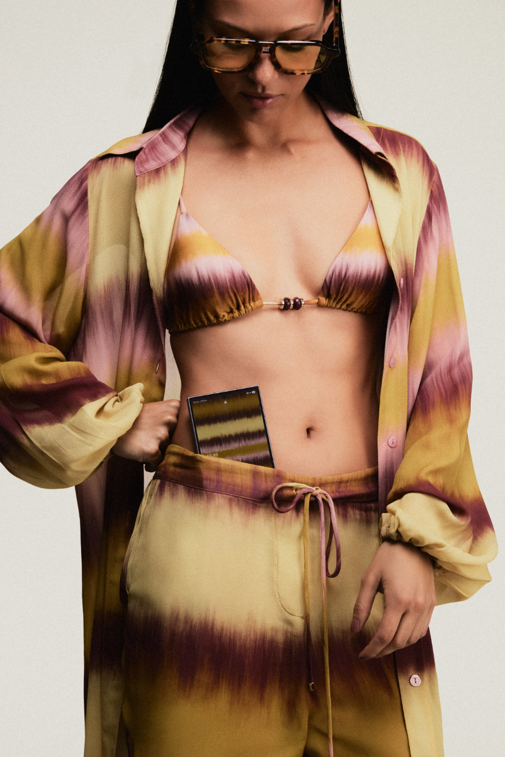 Modelo veste biquíni, camisa e calça com estampa tie-dye da coleção da ÁGUA DE COCO e segura um Galaxy Z Fold8 na cintura.
