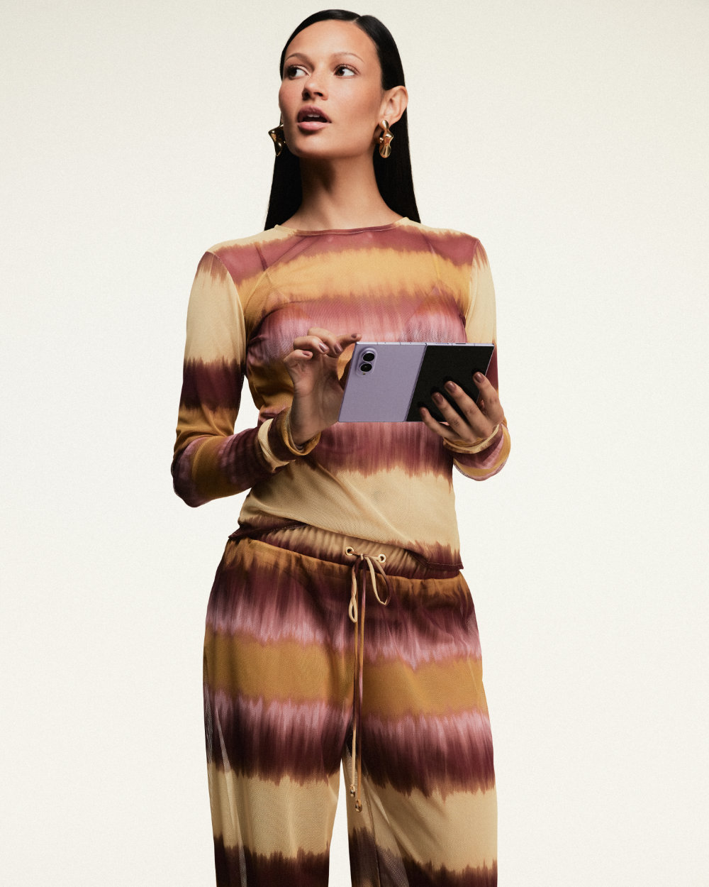 Modelo veste conjunto de blusa de manga longa e calça com estampa tie-dye da coleção da ÁGUA DE COCO e usa um Galaxy Z Fold8 aberto enquanto toca na tela.